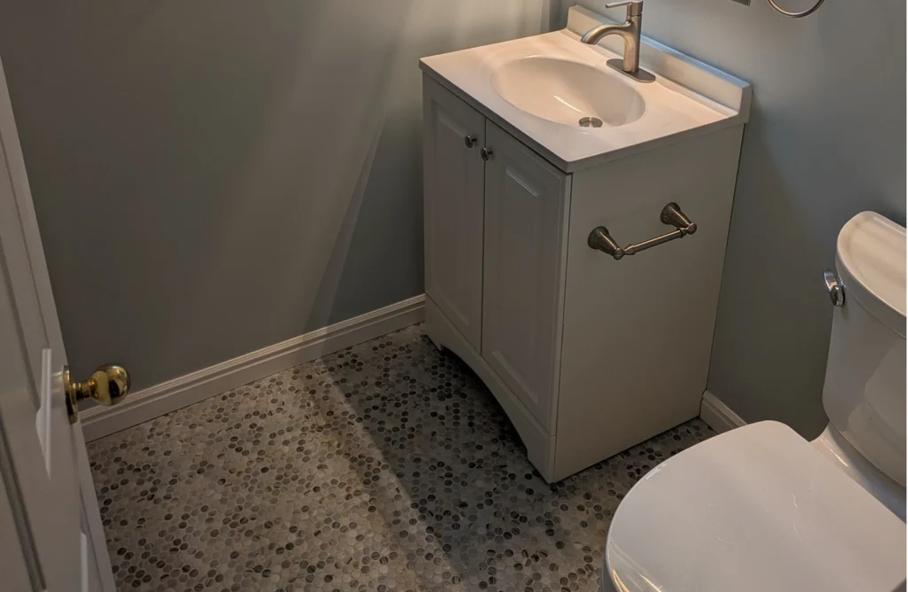A full view of a small bathroom with gray walls, a window, a toilet, and a white vanity with a sink.